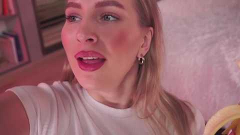 Media: Video of a smiling, fair-skinned woman with blonde hair and pink lipstick, wearing a white top, sitting in a pink room with a bookshelf in the background.