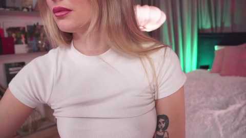 Media: Video of a blonde woman with pink lips, wearing a tight white T-shirt, showing a tattoo on her left arm, in a softly lit bedroom with pink curtains and a fluffy white bedspread.