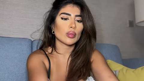 Media: Video of a young woman with long dark hair, light skin, and full lips, wearing black lingerie, sitting on a blue couch in a modern living room.