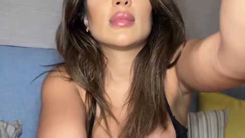 Media: Video of a young woman with long, straight brown hair, light skin, wearing a black bra. She's puckering her lips in a kiss. Background shows a blue wall and a yellow pillow.