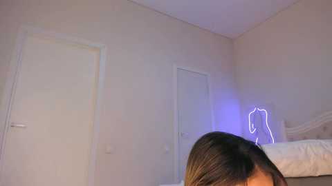 Media: A video of a woman's back in a dimly lit, minimalist bedroom with beige walls and white doors. She has long, dark hair and a neon light in the shape of a heart behind her.