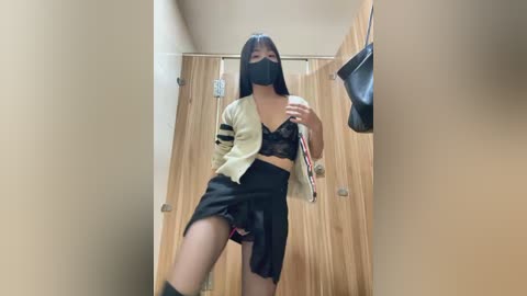 Media: Video of a young Asian woman in a dressing room, wearing a black mask, a white jacket, black lace bra, and high-waisted shorts, holding a red and black bag, with wooden walls and hanging clothes in the background.