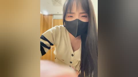 Media: Video of an East Asian woman with long black hair, wearing a beige cardigan, black face mask, and a white top, standing in a wooden room with a beige wall and a ceiling fan.