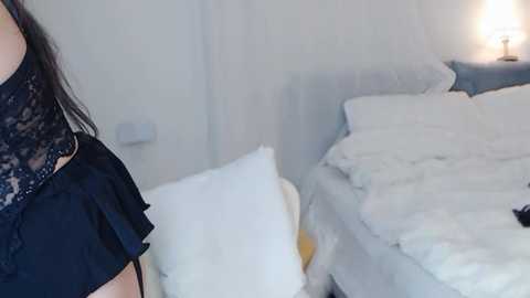 Media: Video of a woman in a black lace top, standing next to a messy, unmade bed with white sheets, in a bright, minimalist room with white walls and a lamp on a nightstand.