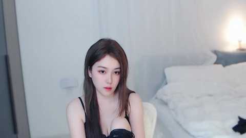 Media: A video of an East Asian woman with long, straight black hair and fair skin, wearing a black lace bra, sitting on a white bed in a minimalist bedroom.