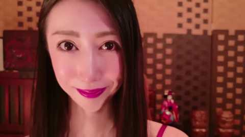 Media: Video of an Asian woman with straight black hair and fair skin, wearing red lipstick, standing in front of a patterned wall with a doll in traditional attire.