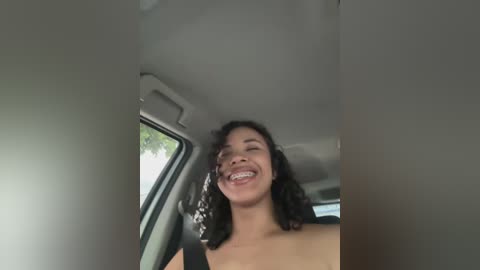 Media: Video of a joyful young woman with medium skin tone and curly black hair, wearing a black top, smiling widely with braces visible. She is seated in a car, looking out the window.