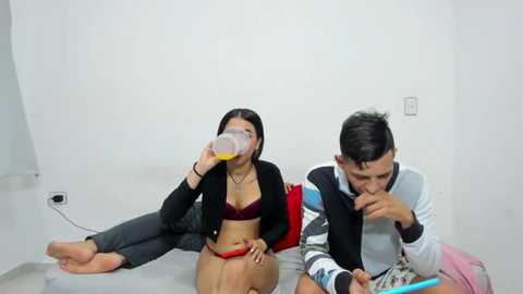 Media: Video of a young woman with long black hair in a red bra and black cardigan, drinking orange juice, and a young man with short dark hair in a striped shirt, eating cereal, sitting on a gray floor in a white room.