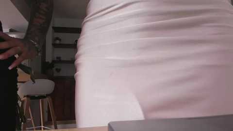 Media: Video of a woman in a tight, white dress, highlighting her curvy figure, with a tattooed arm visible. Background shows a modern, minimalist living room with wooden furniture and white walls.