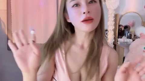 Media: A video of a young Asian woman with long, straight hair, fair skin, and full lips, wearing a low-cut, light pink top, gesturing with her hands. Background features a mirror, fluffy pillows, and a wooden wardrobe.