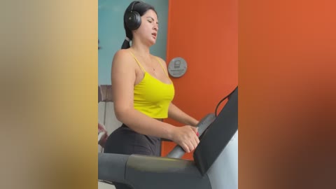 Media: A video of a fit woman with medium skin tone, wearing a tight yellow sports bra and black leggings, using an elliptical machine in a gym with orange and blue walls.