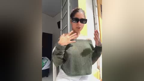 Media: A candid video of a woman with dark hair, sunglasses, and an olive-green sweater, standing in a hallway, making a \"stop\" gesture with her hands.