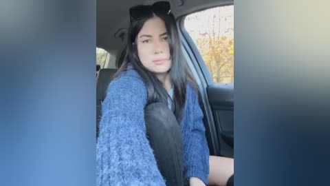 Media: Video of a young woman with long black hair, wearing a blue knit sweater, black sunglasses, and a black seatbelt, seated in a car with autumn leaves visible outside.