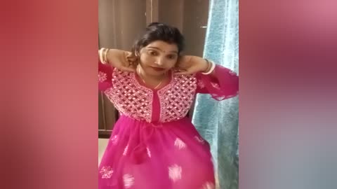 Media: A video of a South Asian woman in a bright pink, embroidered kurta, standing in a bathroom with a teal towel and brown wooden walls.