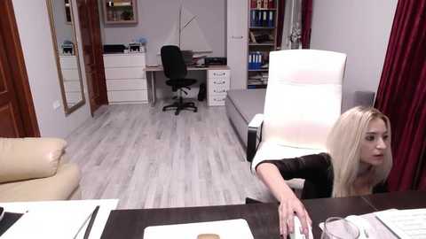 Media: Video of a tidy, modern office with a blonde woman in a black top typing at a desk, surrounded by white walls, a white chair, and wooden floor.