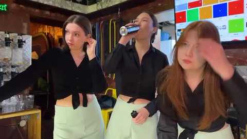 Media: Video of three young women in a cluttered store, wearing black tops and light green pants, with one drinking from a can, another holding a phone to her ear, and the third covering her face.