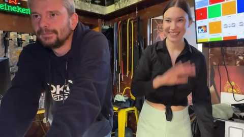 Media: Video of a man with short gray hair and beard in a dark hoodie, and a woman with brown hair tied back, wearing a black blouse and light skirt, in a cluttered, brightly lit room with a TV screen displaying a colorful graphic.