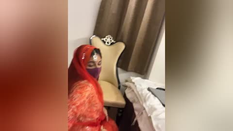 Media: Video of a woman in a red sari and purple mask, sitting on a beige chair with a decorative backrest. The background features a white wall and brown curtains. The image is slightly blurred and has a reddish tint.