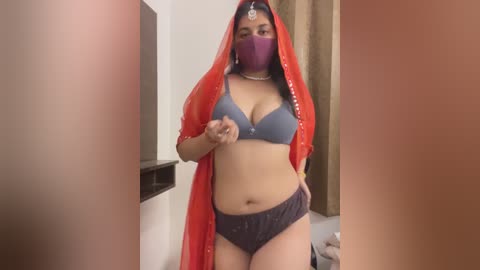 Media: Video of a curvy South Asian woman wearing a red veil, purple face mask, and blue bra, standing in a dimly-lit room.