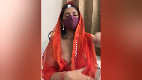 Media: Video of a South Asian woman with medium skin tone, wearing a sheer red sari, purple face mask, and a white pearl necklace, sitting with eyes closed, against a beige curtain background.