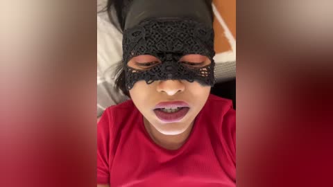 Media: Video of a person with medium skin tone, wearing a black lace mask and red lipstick, lying on a bed with white sheets, wearing a red shirt.