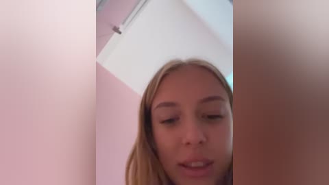 Media: Video of a young Caucasian woman with light skin and straight blonde hair, slightly smiling, taken from a low angle, with a blurred background of white and pink walls.