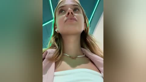 Media: Video of a young Caucasian woman with light skin and blonde hair, wearing a white off-shoulder top and light pink jacket, under neon green ceiling lights.