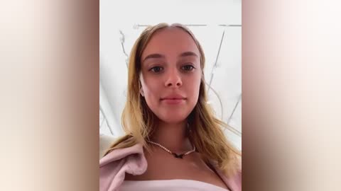 Media: A video of a young Caucasian woman with long, light brown hair, wearing a white top and a pink cardigan, standing in a white, minimalist room with a blurred background.