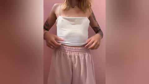 Media: Video of a light-skinned woman with blonde hair, wearing a white tank top and loose, light pink pajama pants, adjusting her waistband. She has tattoos on her arms. Background is a soft pink wall.