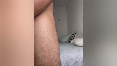 Media: Video of a nude man's thigh and butt in a minimalist bedroom with a white bed and pillows, featuring a light-colored sheet.