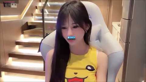 Media: Video of a young Asian woman with long black hair wearing a Pikachu tank top, gagged with a blue duct tape over her mouth, sitting in a white gaming chair in a modern, well-lit room with wooden stairs and a fridge in the background.