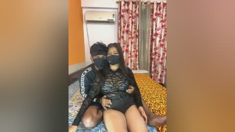 Media: A video of an Asian couple in black face masks, seated on a bed with leopard print sheets, in a room with orange walls, red floral curtains, and an air conditioning unit.