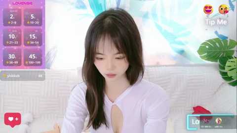 Media: Video of a young Asian woman with long black hair, wearing a white blouse, sitting indoors with a vibrant blue background. Virtual chat interface overlays show her stats and user information.