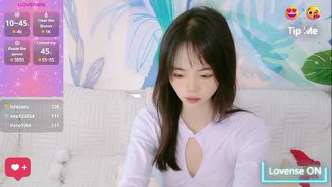 Media: Video of an Asian woman with long black hair, wearing a light purple top with a keyhole cutout, sitting on a white couch with a colorful, abstract painting behind her. She appears to be looking at her phone.