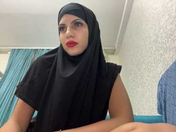 Media: Video of a South Asian woman with fair skin, wearing a black hijab and matching dress, with bold red lipstick, sitting in a hospital room with white walls, teal curtains, and a blue bedspread.