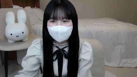 Media: Video of an East Asian woman with long black hair, wearing a white face mask, gray sweater, and a black bow tie, sitting on a white bed with a stuffed bunny beside her.