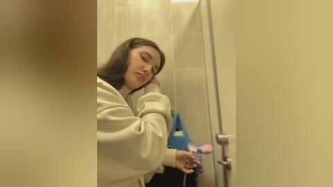 Media: Video of a young woman with long dark hair in a beige sweater, leaning against a bathroom stall door, looking contemplative. The background shows tiled walls and a blue bag on the floor.