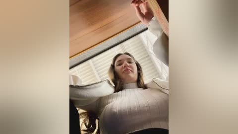 Media: A video of a young woman with shoulder-length brown hair, wearing a white, ribbed sweater, standing near a window with closed blinds. Her expression is neutral, and the image has a soft, blurred effect.