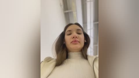 Media: Video of a young woman with long brown hair, wearing a cream-colored turtleneck sweater, standing against a blurred white background with a window. Her expression is neutral.