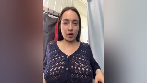 Media: Video of a young woman with light skin and long brown hair, wearing a navy crochet top revealing her large breasts, seated in a bathroom with a shower curtain and towel rack in the background.