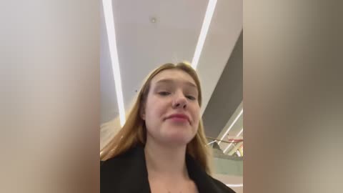 Media: Video of a young woman with fair skin and straight, shoulder-length blonde hair, wearing a black blazer, standing in a modern indoor setting with a white ceiling and recessed lights.