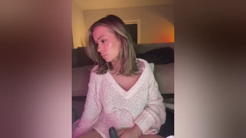 Media: Video of a blonde woman in a beige knit sweater, sitting in a dimly lit living room, with a blurred background featuring a couch and a warm glow from a lamp.