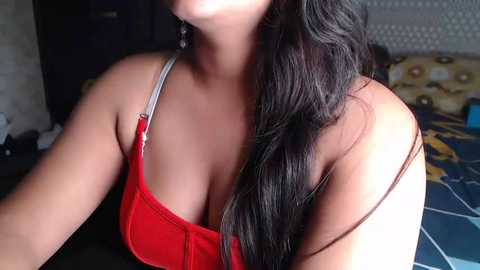 Media: Video of a young woman with long, wavy black hair, wearing a red bra, captured from the chest up, showing a close-up of her cleavage and shoulders in a dimly lit room with a patterned bedspread in the background.