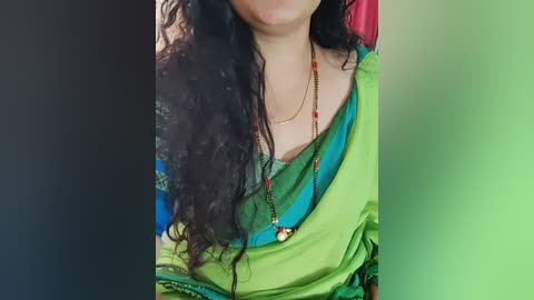 Media: Video of a woman with long, curly dark hair wearing a green and blue sari, gold necklace, and a red beaded bracelet.