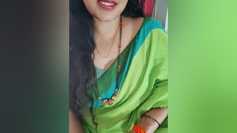 Media: Video of a woman with long black hair, wearing a green sari with turquoise accents, beaded necklace, and orange wristband, smiling. Background features a blurred green and black gradient.