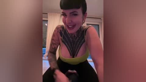 Media: Video of a tattooed woman with short black hair, wearing a yellow tank top and black pants, smiling, leaning against a wall with a window behind her.