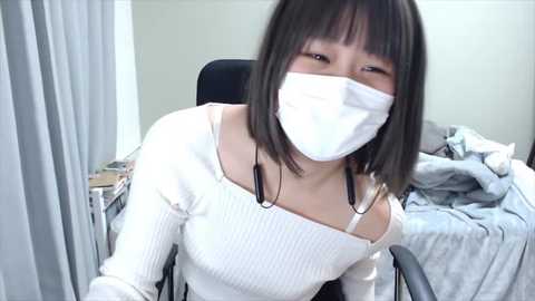 Media: Video of an East Asian woman with short brown hair, wearing a white face mask, white ribbed sweater, and black headphones, seated in a chair in a cluttered, dimly lit room with a gray bed and scattered clothes.