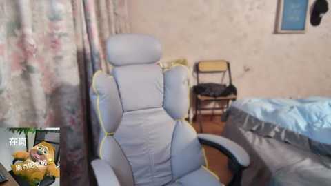 Media: Video of a beige gaming chair in a cluttered bedroom with floral curtains, a messy bed, wooden chair, and a small table with a yellow chair.