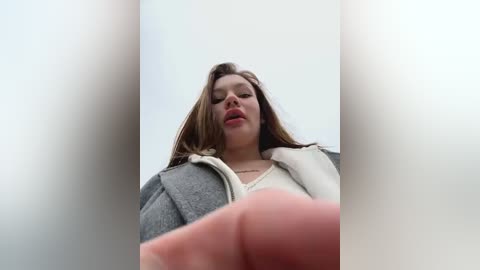 Media: A video of a young woman with light skin and brown hair, wearing a grey coat and white scarf, looking upward with a slightly open mouth, in a blurry, minimalist background.