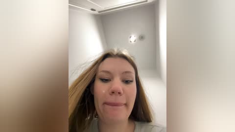 Media: Video of a young woman with light skin, long straight blonde hair, and a neutral expression, taken from a low angle, showing her from the chest up. The background features a plain, white wall with a small, round light fixture.